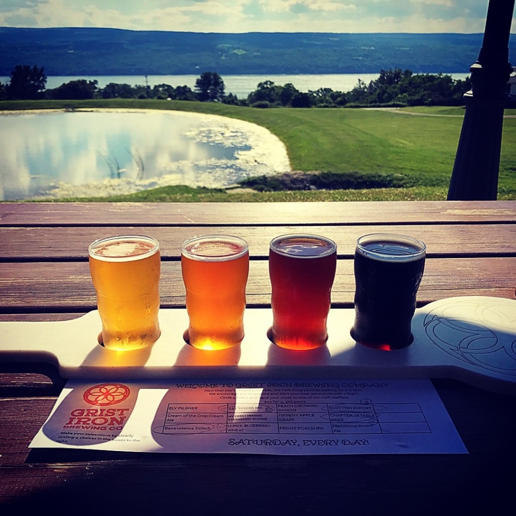 Small taster glasses of beer arranged from light to dark on a wooden serving paddle.  View of a reflective pond and a lake in the background.